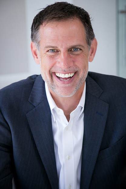 Man in navy blazer and white shirt smiles widely in bright office setting.