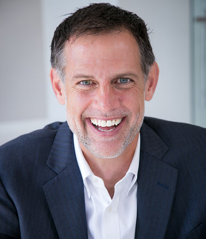 Man in a navy blazer and white shirt smiles broadly, indoors.