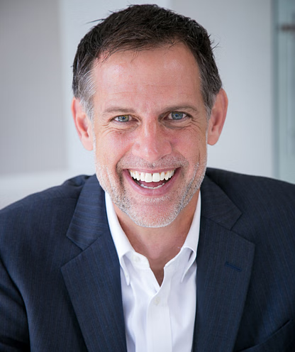 Man in a navy blazer and white shirt smiles broadly, indoors.
