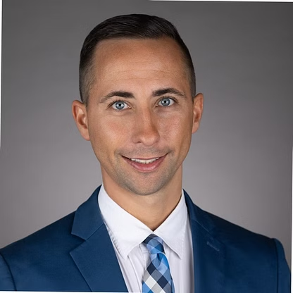 Man in a blue suit smiles, blue patterned tie, against gray backdrop.