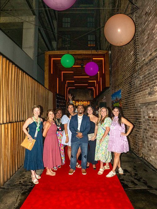 Group standing on a red carpet in a narrow brick alley with hanging balloons and warm lights