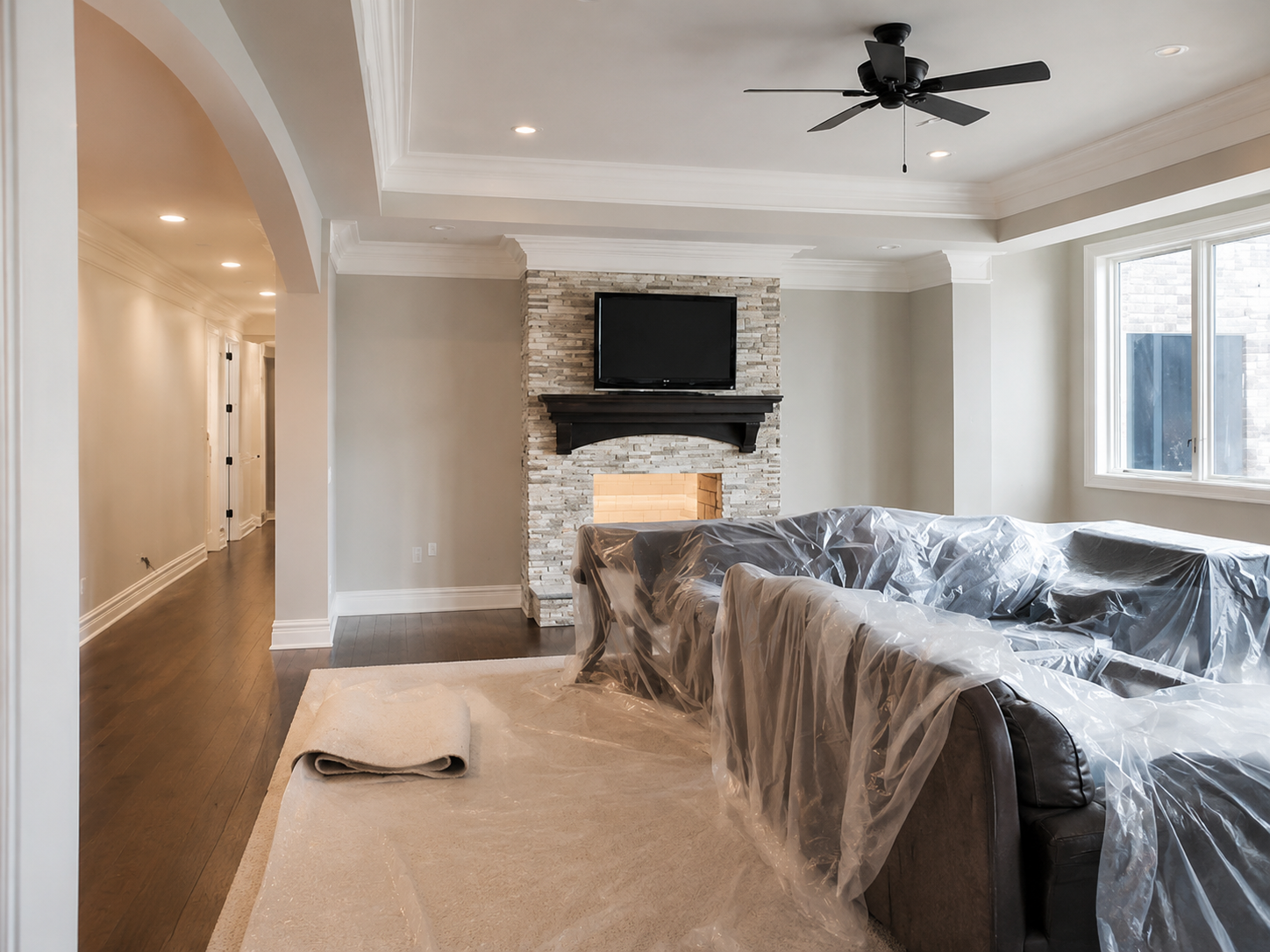Living room under renovation, with plastic-covered furniture, fireplace, ceiling fan, and large windows