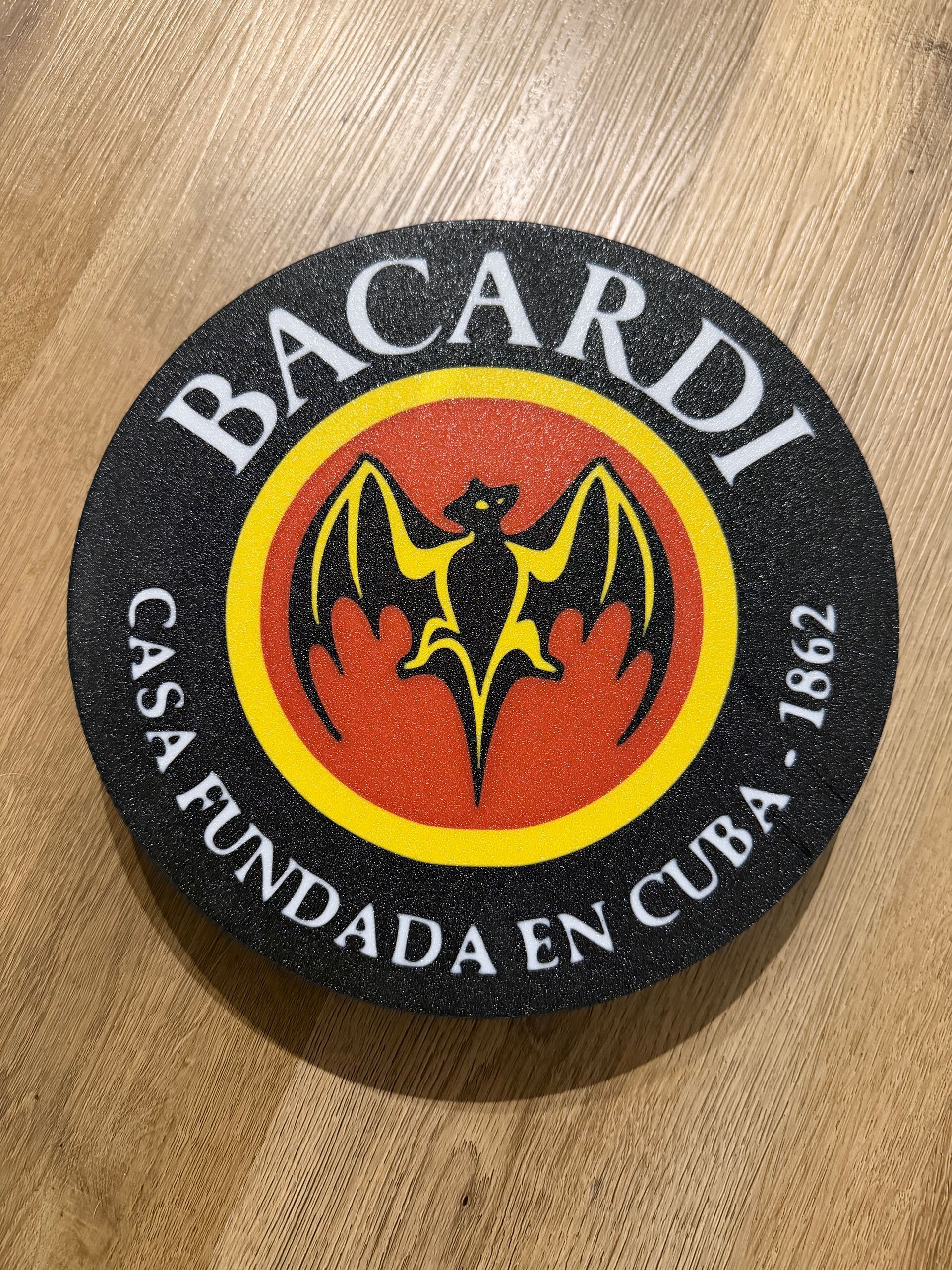 Bacardi-onderzetter met zwarte rand, rood middenstuk en geel vleermuislogo op een houten ondergrond.