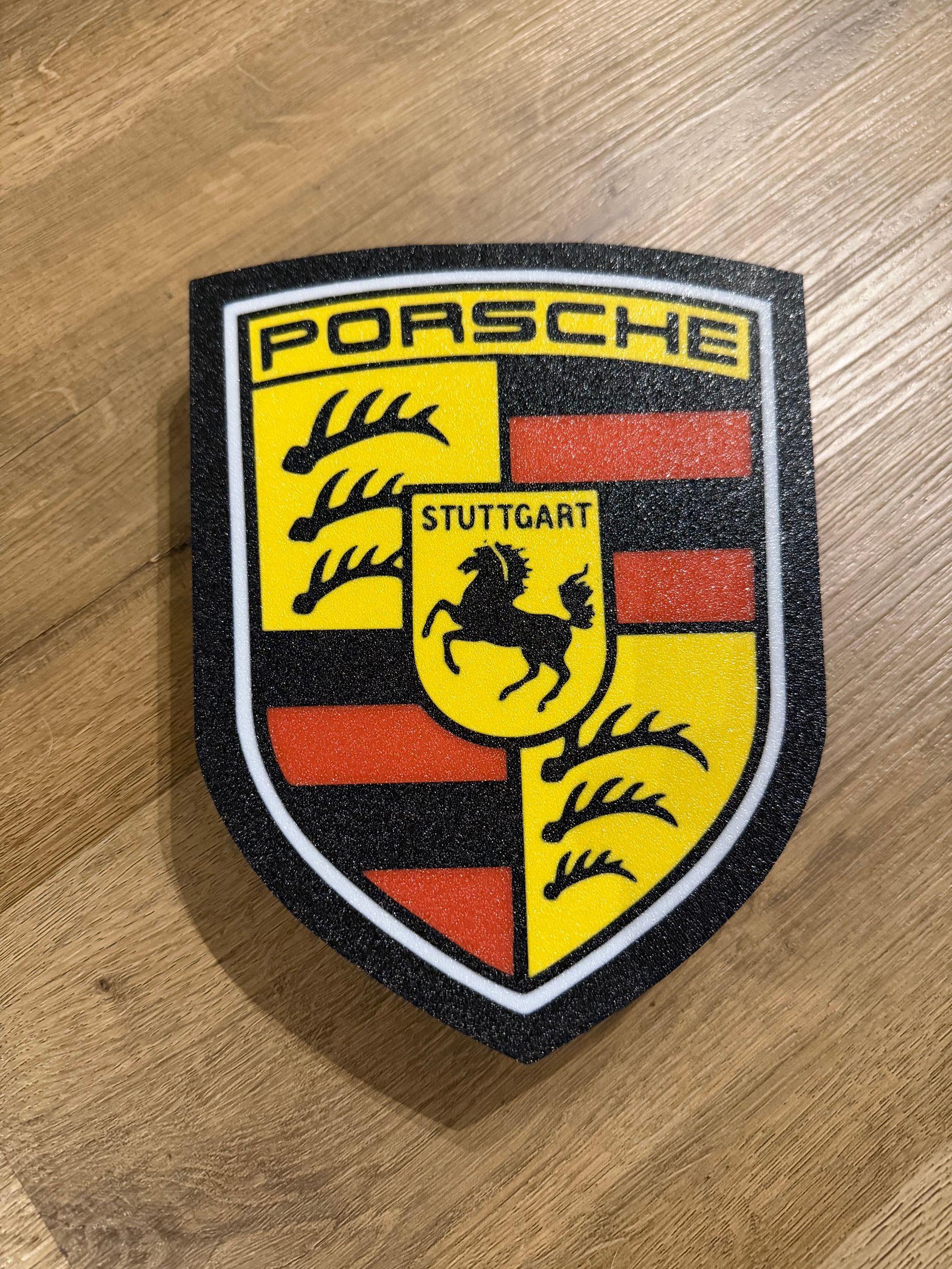 Porsche-schildlogo op een houten ondergrond, met gele, zwarte, rode en zilveren details.