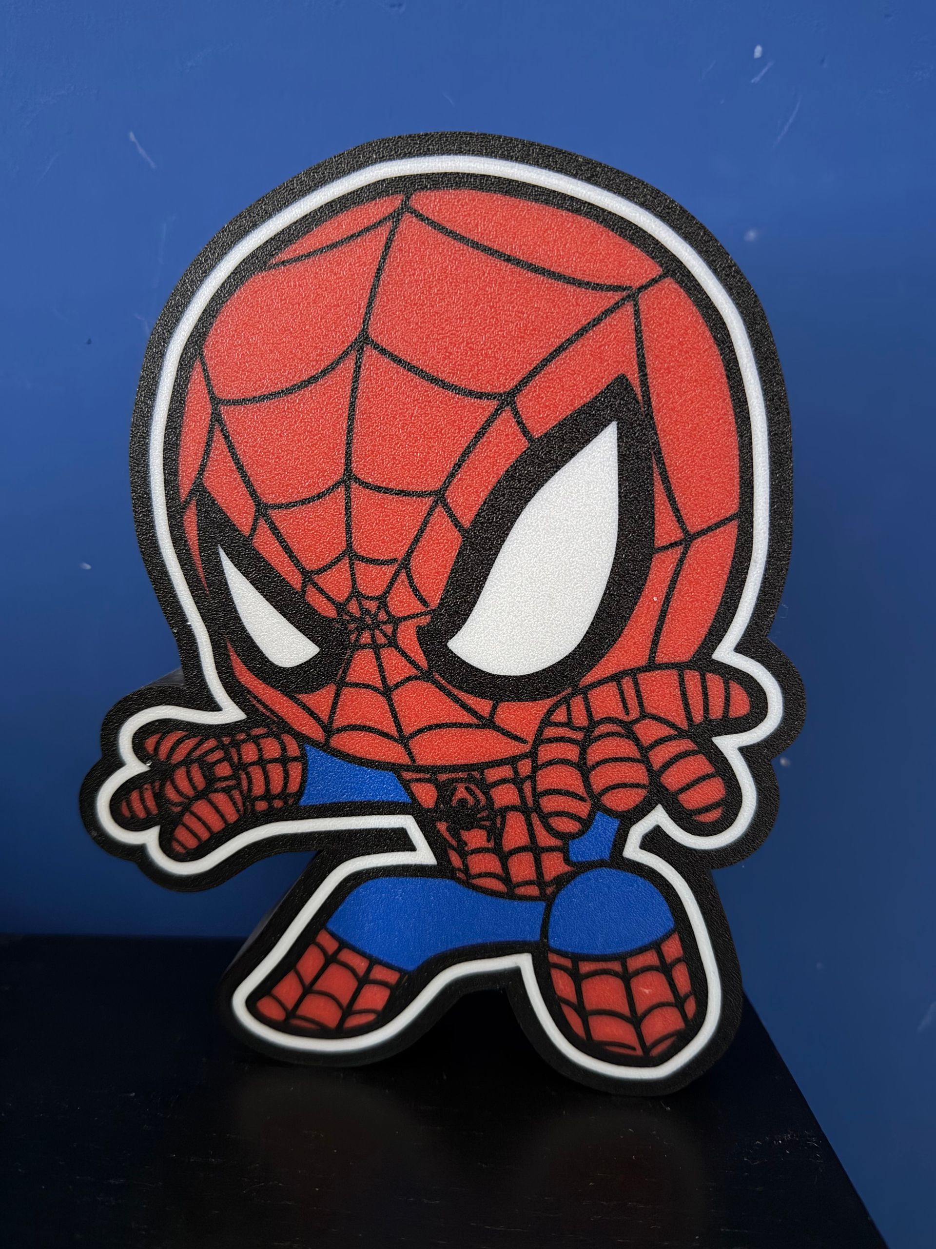 Pluche Spider-Man-figuurtje met grote ogen, rood webmasker en blauwe broek tegen een blauwe achtergrond.