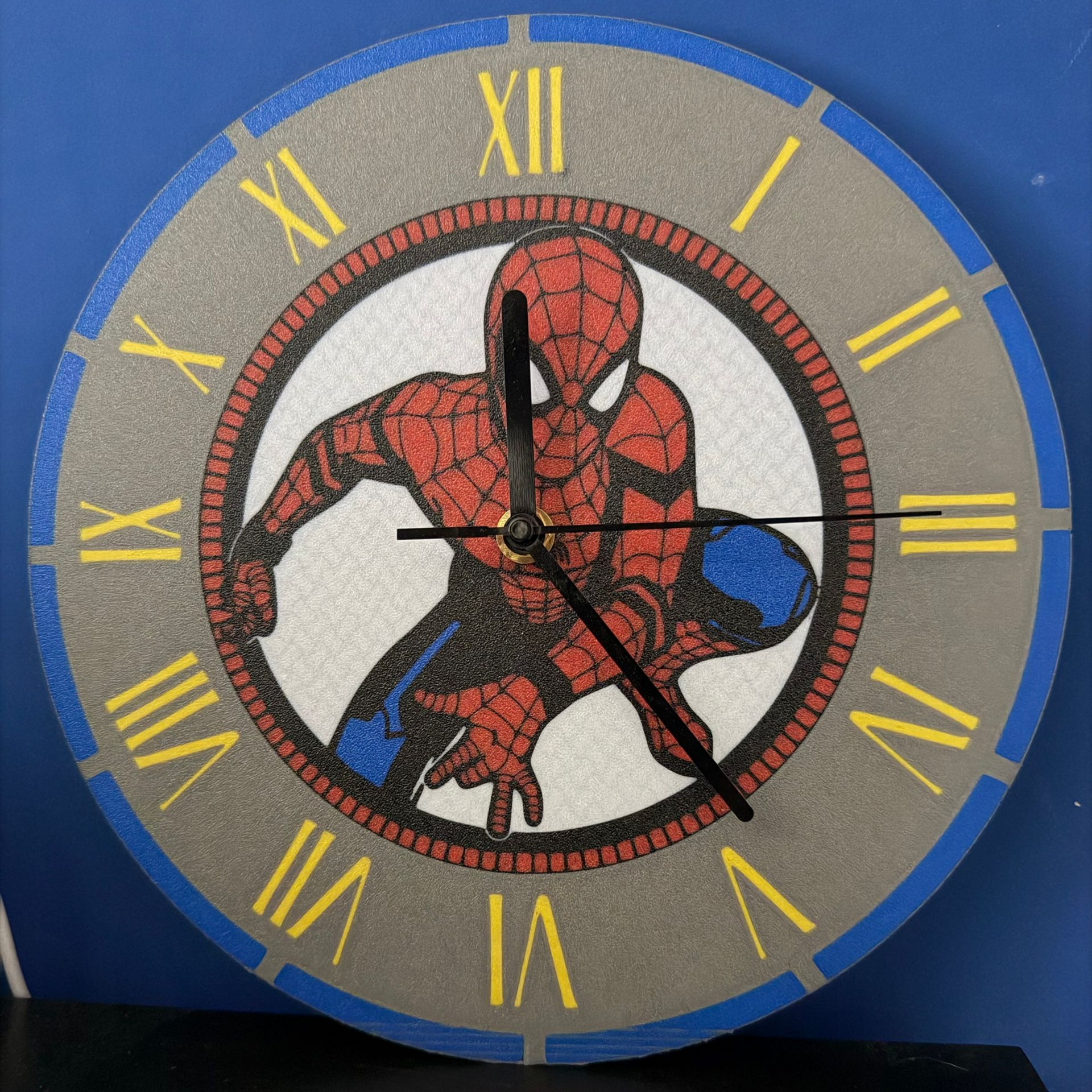 Spider-Man op een blauwe wandklok met gele Romeinse cijfers en zwarte wijzers.