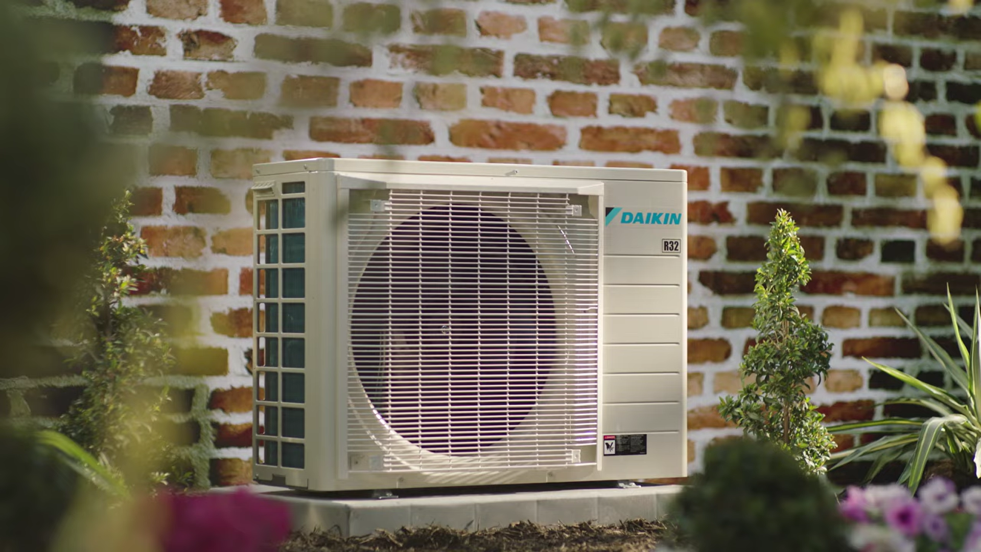 Outdoor air conditioning unit in front of a brick wall, surrounded by plants and sunlight.