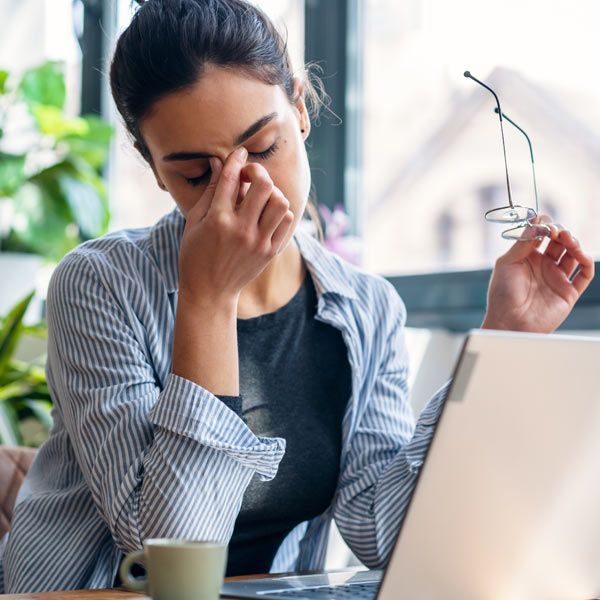 A woman with a bad headache is sitting at a desk with a laptop, rubbing their eyes with one hand and holding glasses in the other.