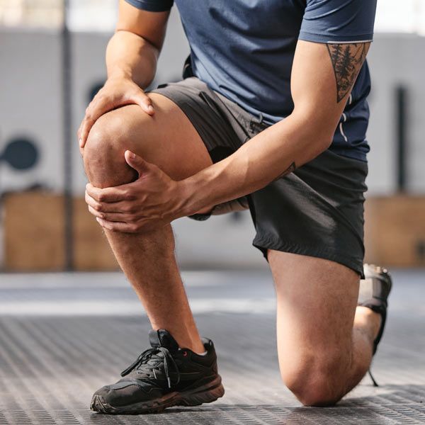 A person in athletic wear is kneeling on one leg in a gym, holding their knee pain with both hands.