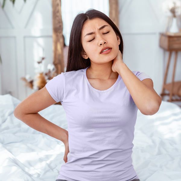 A person sitting on a bed, clutching their neck and lower back in apparent pain with their eyes closed.