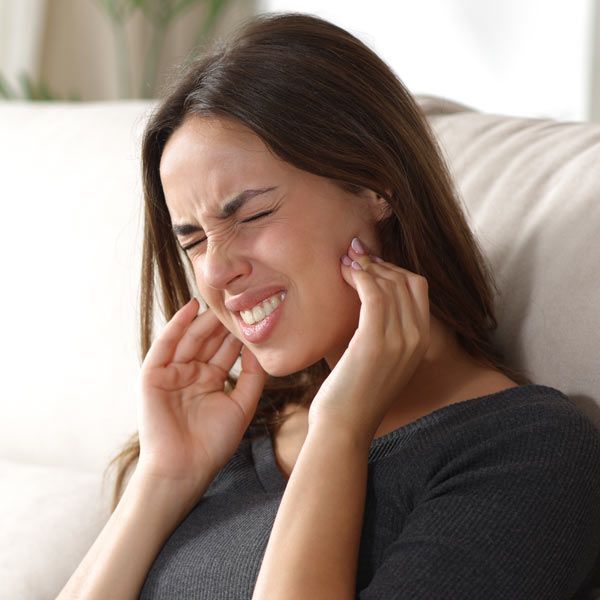 A person with TMJ is sitting on a sofa, grimacing in pain while holding both sides of their jaw with their hands.