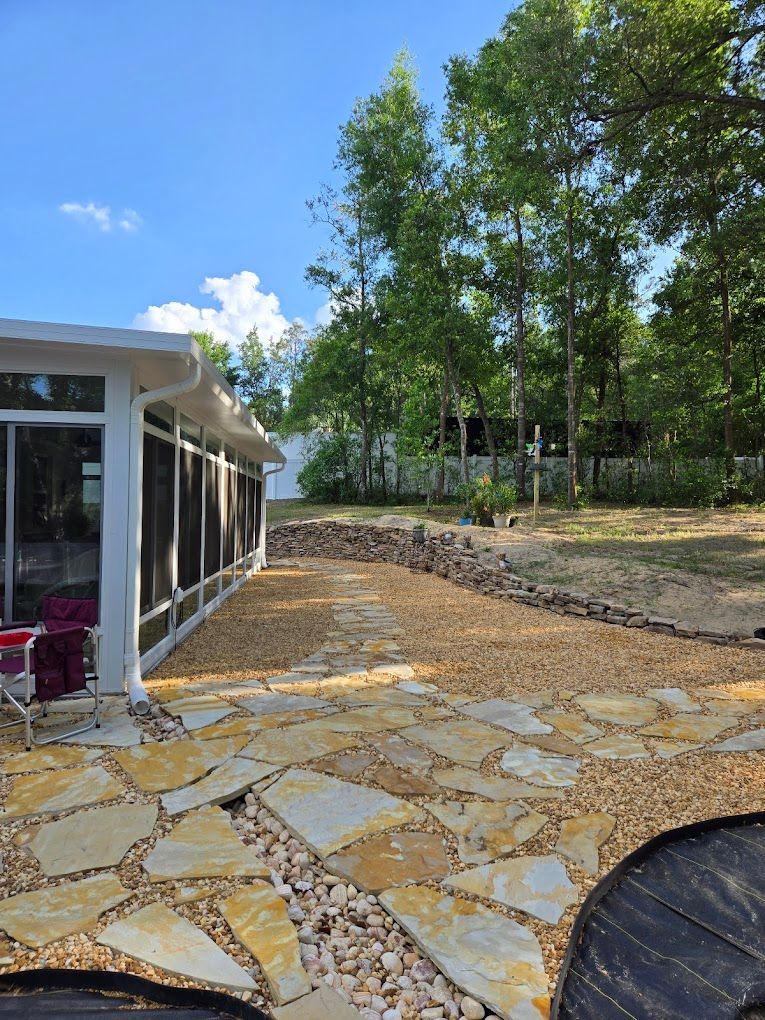 Stone patio next to a sunroom, transitioning to gravel, then a stone retaining wall against a backdrop of trees under a blue sky.
