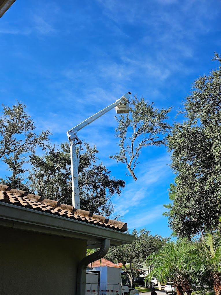 A white boom lift trimming a tree near a roof on a sunny day.