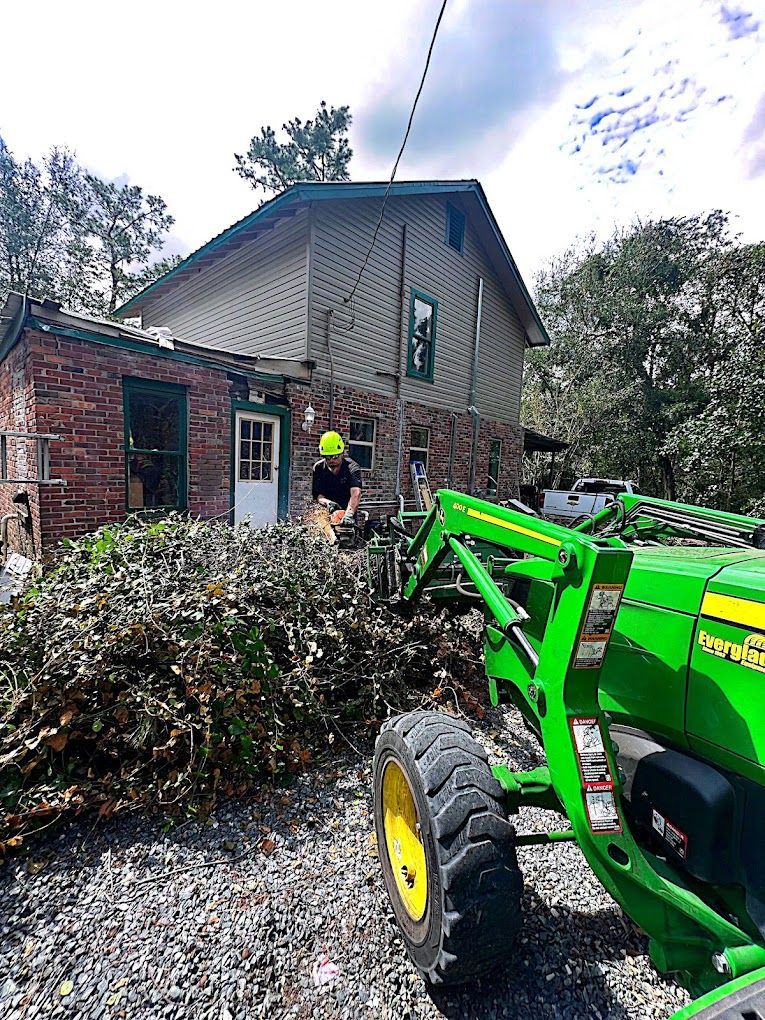 Man using chainsaw to cut branches near a two-story house, a green tractor in the foreground, and a pile of cut limbs.