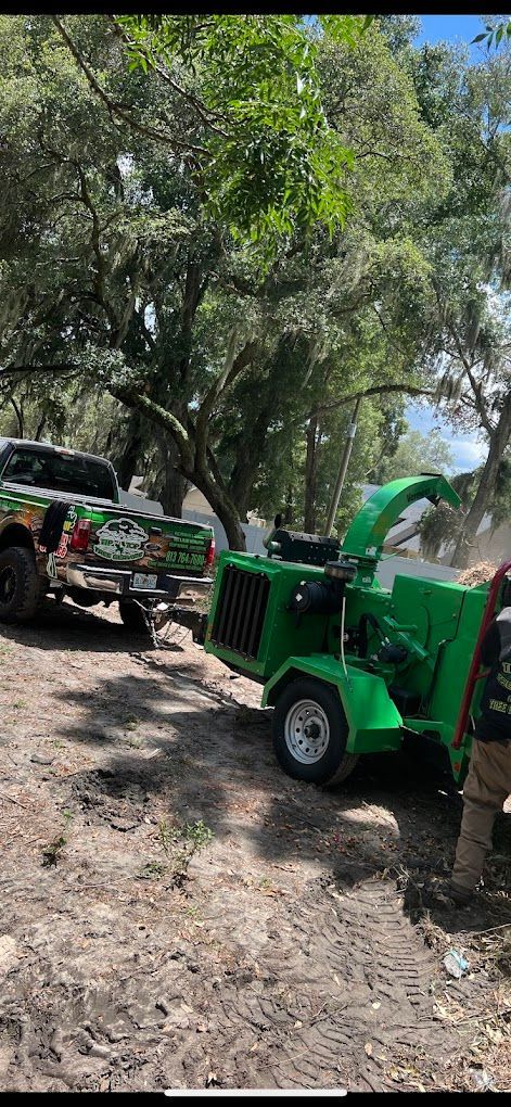 A green wood chipper attached to a truck, in a wooded area.  A person is standing nearby.