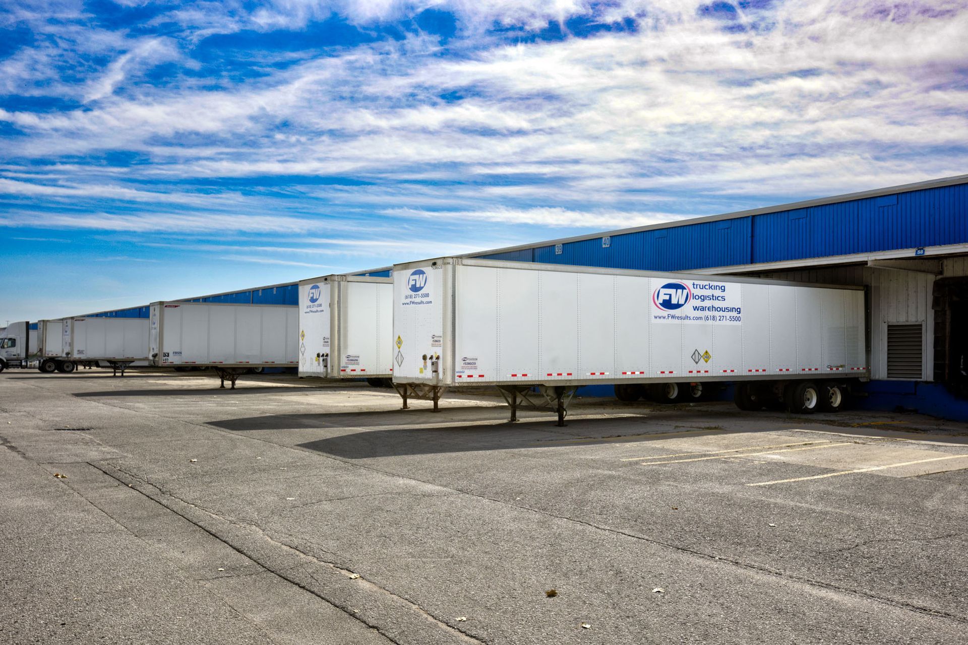 A warehouse with multiple truck trailers backed up to the bays of a warehouse and clouds in sky.