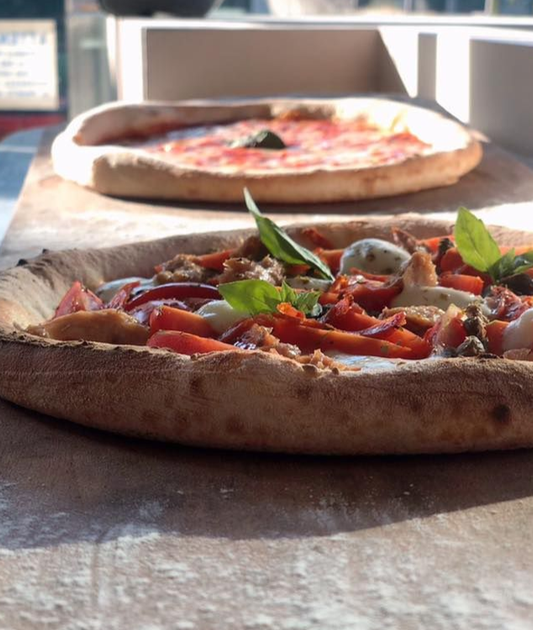 Due pizze su un tavolo, una con pomodori e basilico in primo piano, l'altra semplice sullo sfondo.