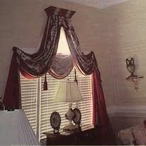 Window with white blinds, burgundy draped curtains, and a lamp on a table in a beige room