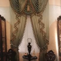 Ornate green drapes framing a window with a lamp and decorative table in a formal room