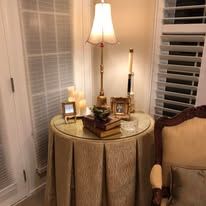Round table with gold skirt, lamp, candles, books, and framed photos beside a beige chair in a cozy corner