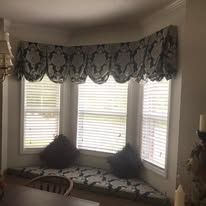 Bay window seat with patterned valance, blinds, and dark throw pillows in a beige room