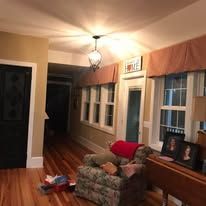 Dimly lit living room with hardwood floors, windows, a patterned chair, couch, and framed photos on a sideboard