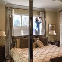 Person opening a window in a beige bedroom with a bed, lamps, and mirrored closet doors