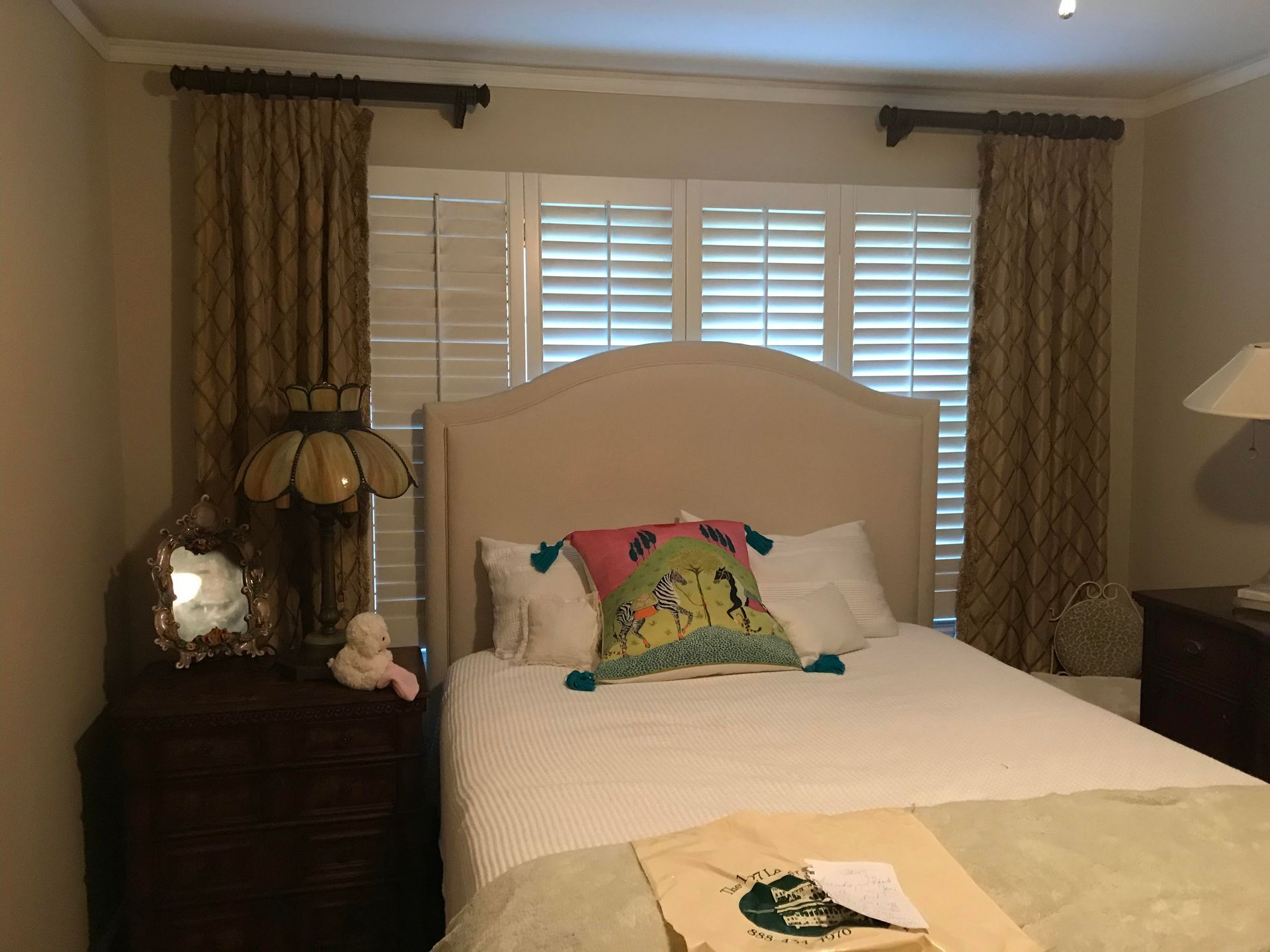 Bedroom with neatly made bed, beige headboard, patterned curtains, and bedside lamps
