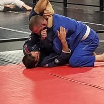 Two wrestlers grappling on a mat, one in a blue gi on top and one in a black gi below.