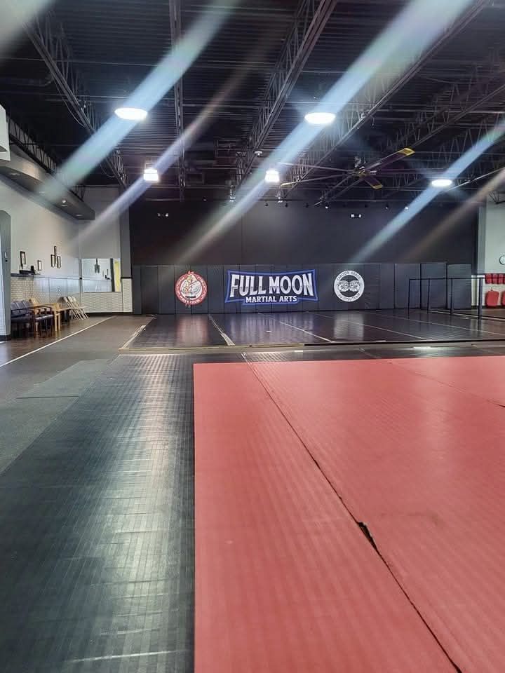 Indoor martial arts gym with red mats, black flooring, and a “Full Moon” banner on the back wall.