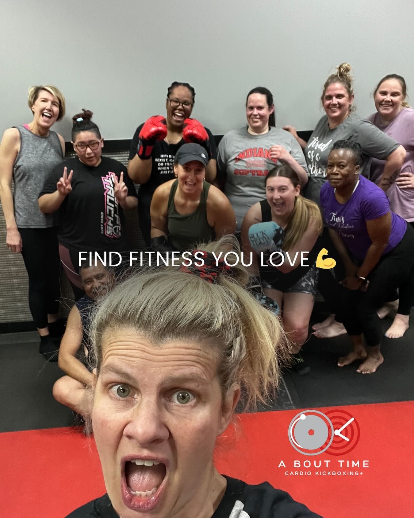 Group of smiling fitness class participants posing in a gym, with one woman making a playful face in front.