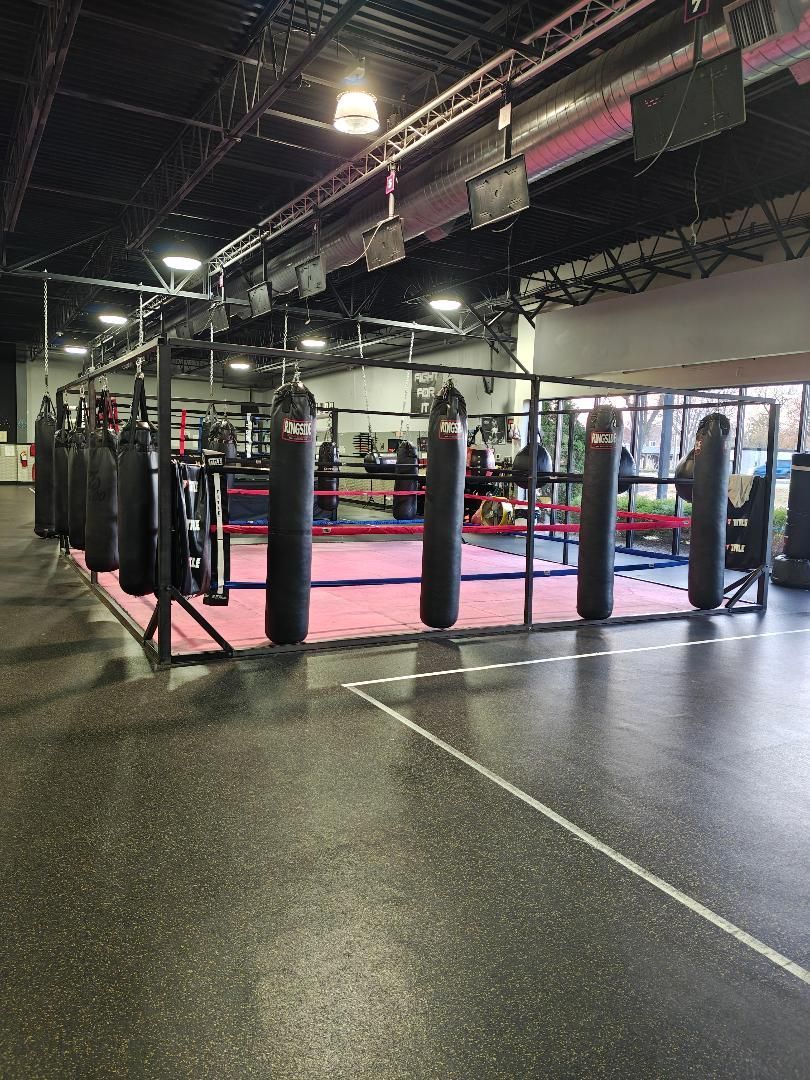 Indoor boxing gym with a pink ring, black punching bags, and a dark speckled floor under overhead lights