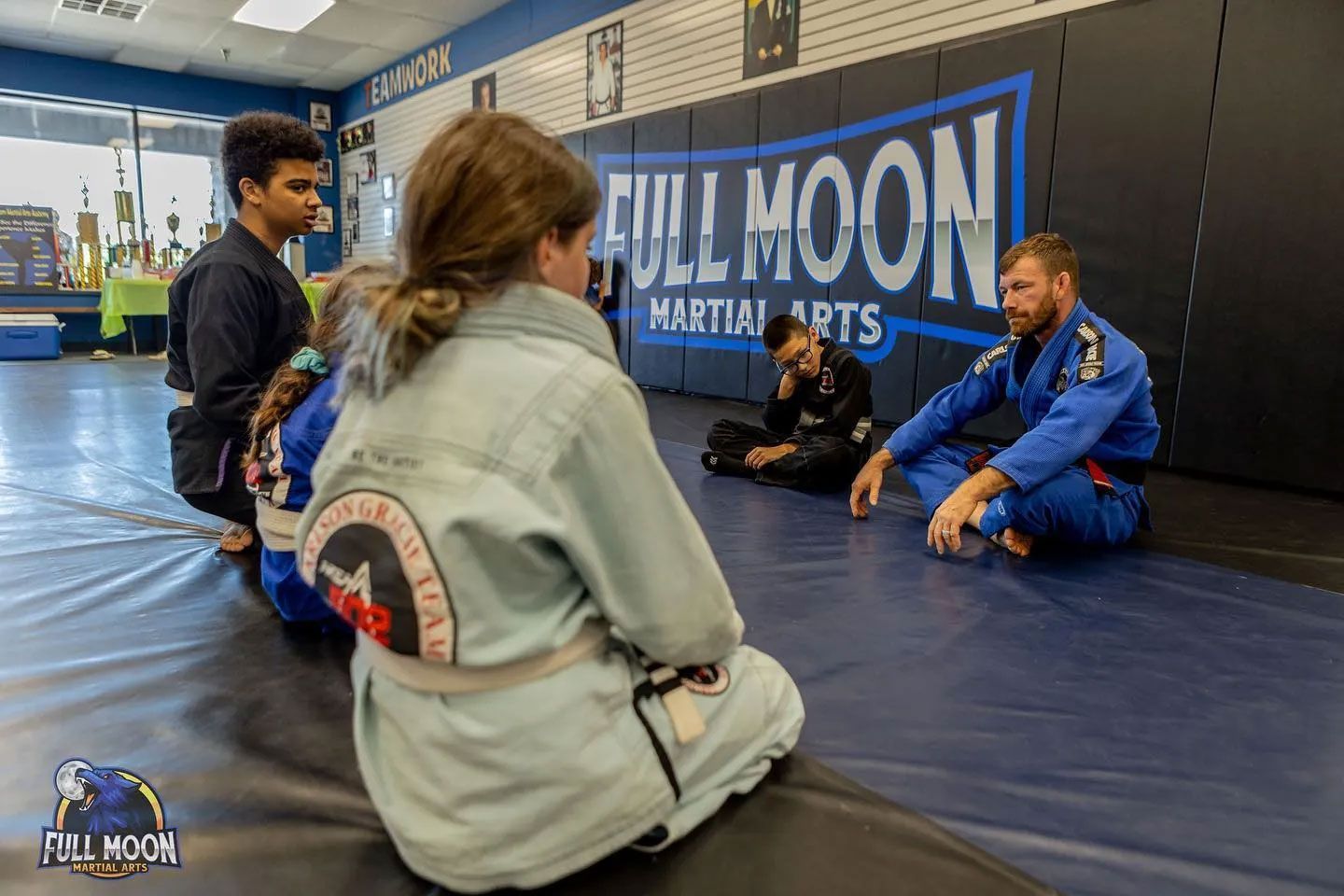 People in martial arts uniforms on a mat, listening to an instructor. 