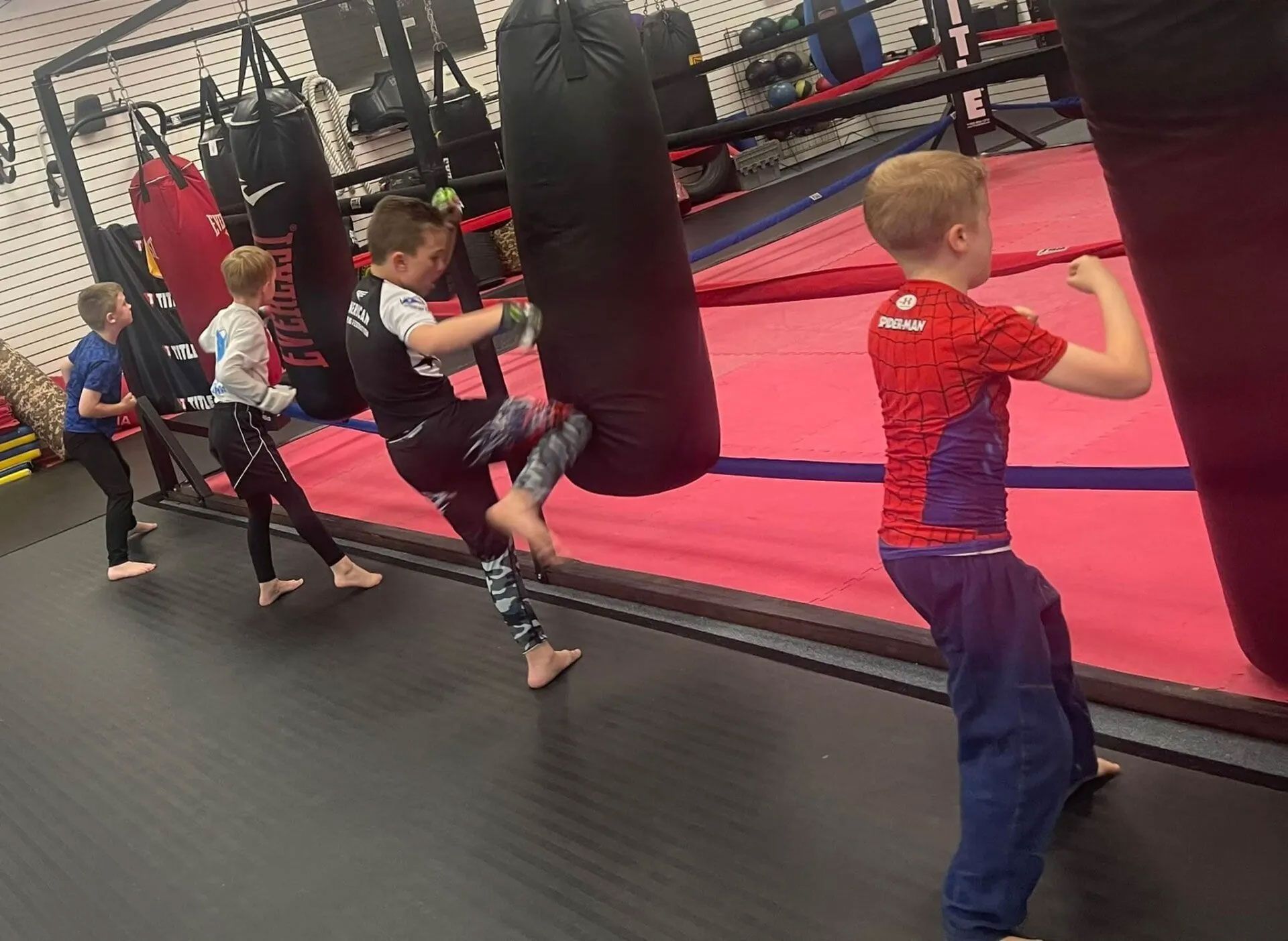 Children practice kicking a punching bag in a gym with red and black equipment.