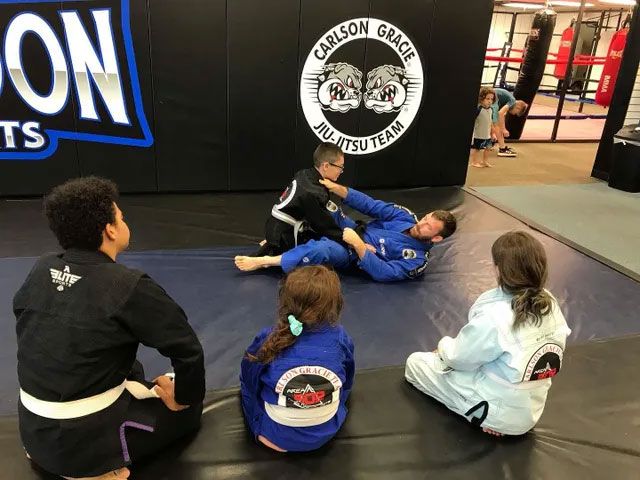 Children in a jiu-jitsu class watching a demonstration. The instructor is on the mat with a student.