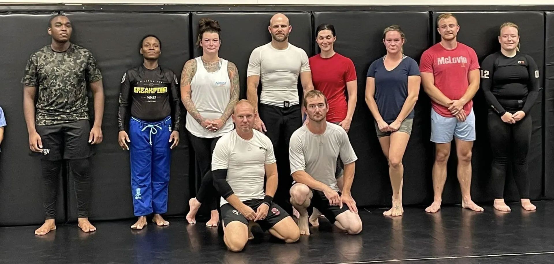 Group of people posing together on a wrestling mat; some are kneeling.