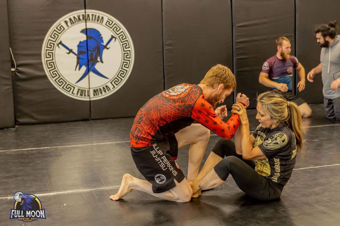 Man kneeling, arm wrestling with a woman in a martial arts training setting.