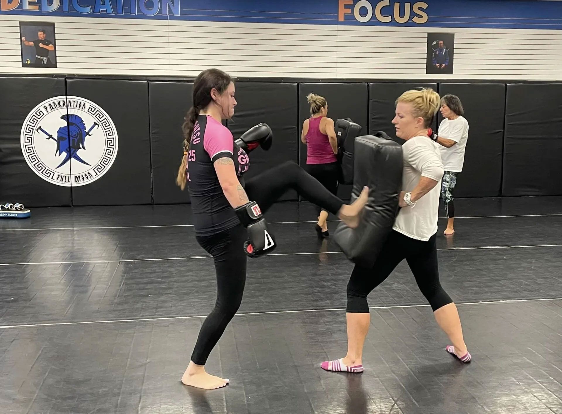 Woman kicking training pad held by another woman in a gym. Other people are in the background.