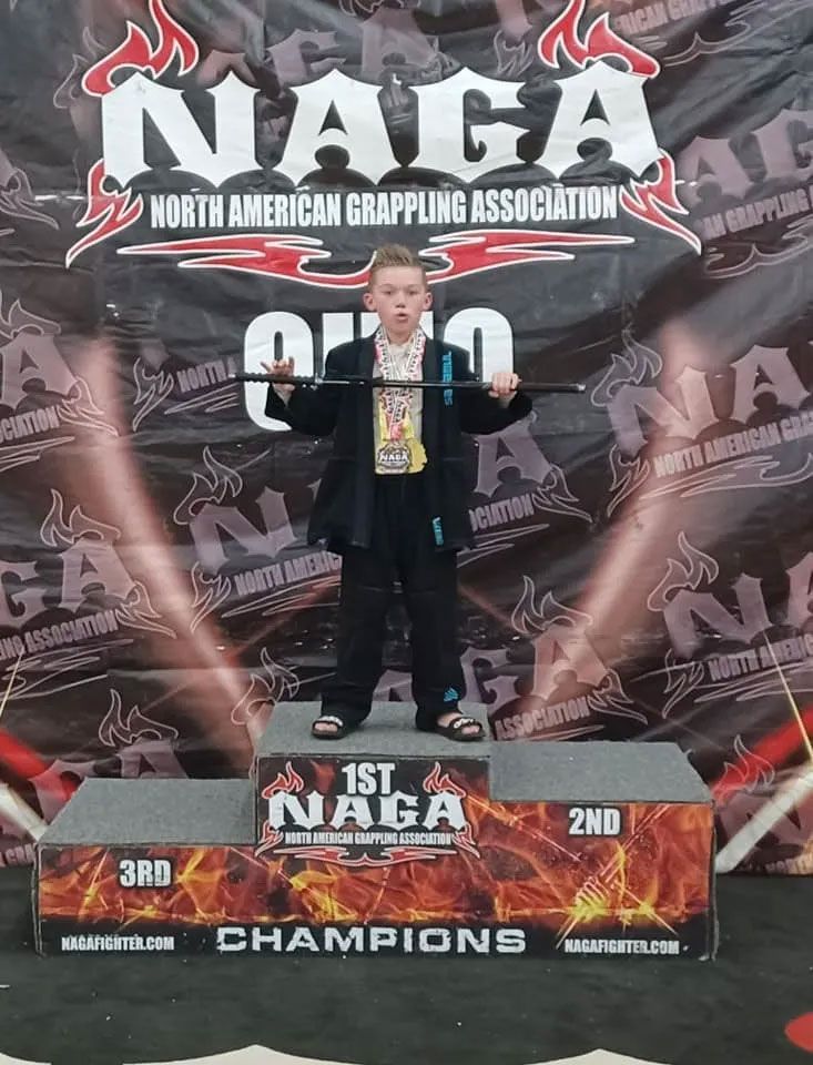 Young person in a suit stands on a podium, holding a trophy, at a NAGA grappling event in Ohio.