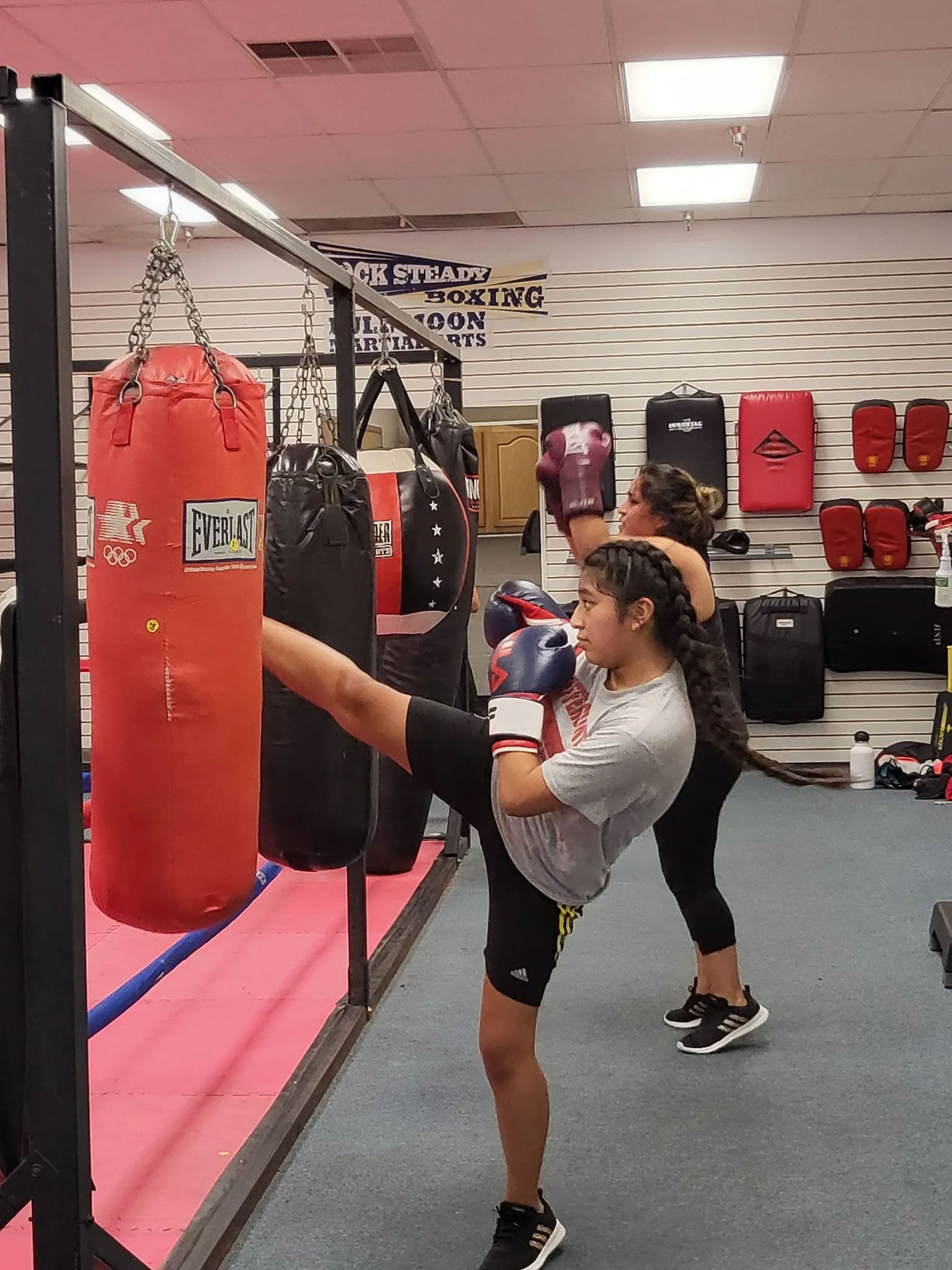 Two people kickboxing a punching bag in a gym. A person in a gray shirt kicks the bag, others punch.