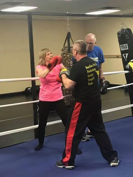 Woman in pink shirt boxing in a ring with a trainer; another man watches.