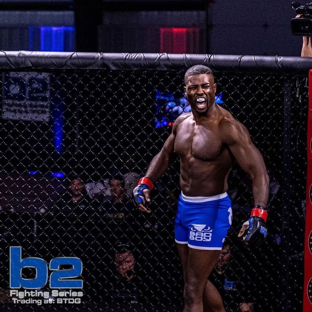 MMA fighter in blue shorts, stands in a cage, smiling with arms raised.