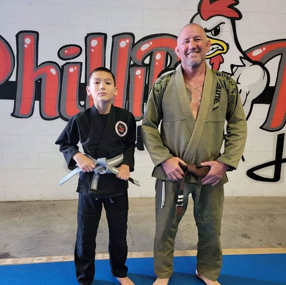 Man and child in martial arts uniforms, standing in front of a gym mural.
