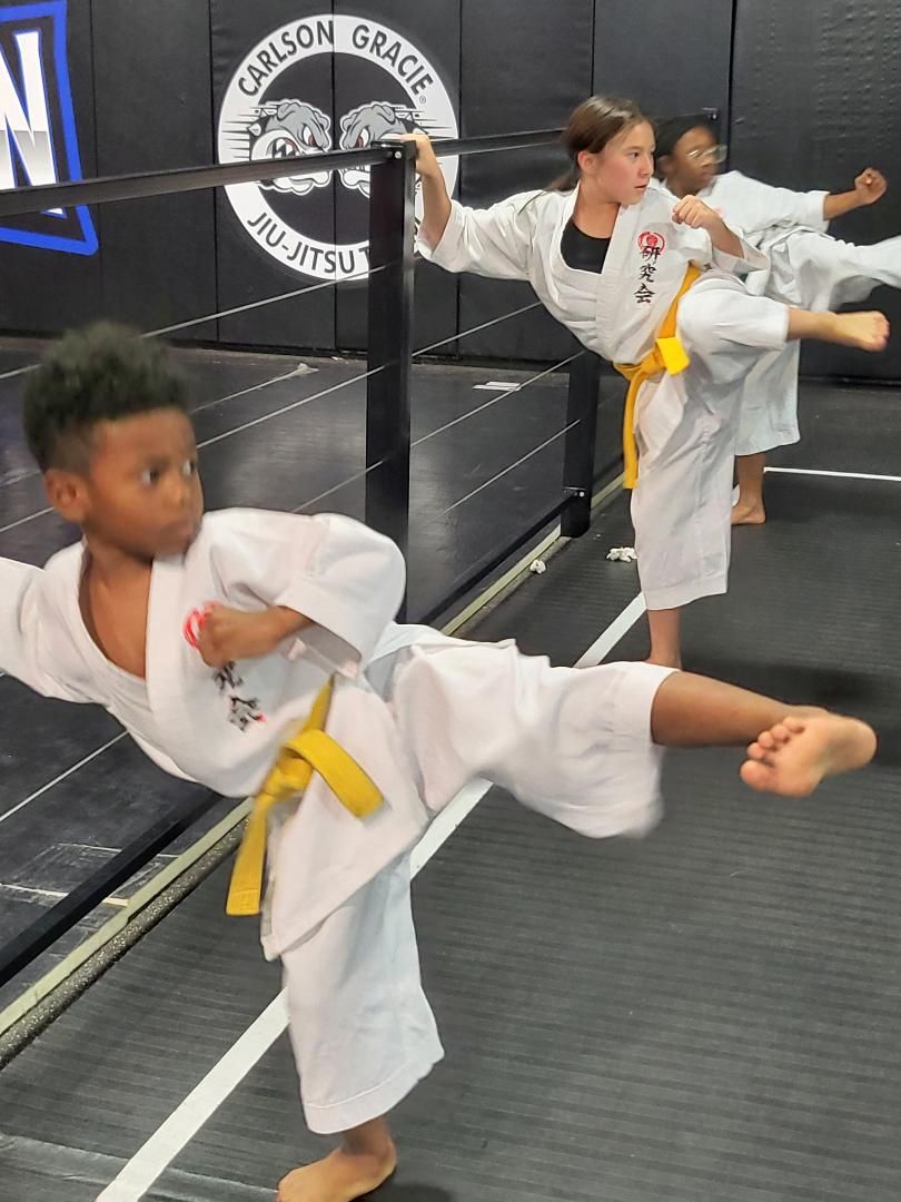 Children in karate uniforms practice kicking and punching in a dojo with a black and blue sign.