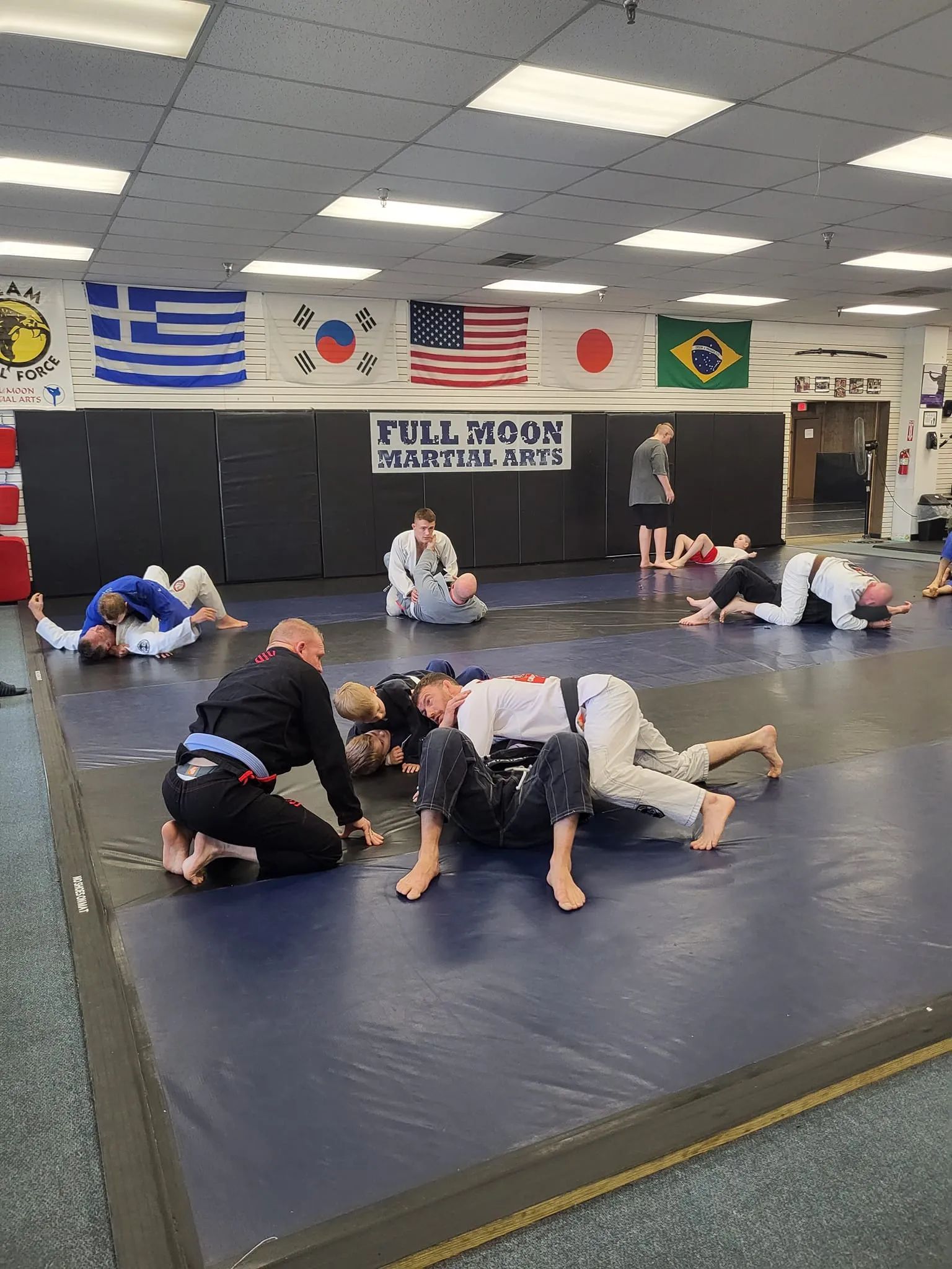 People practicing Jiu-Jitsu on mats in a gym, wearing different colored belts, some are grappling while others watch.