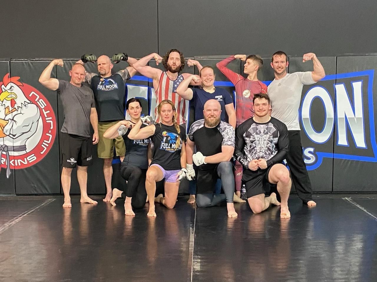 Group of people flexing biceps in a gym. Mats on the floor, wall logos visible.