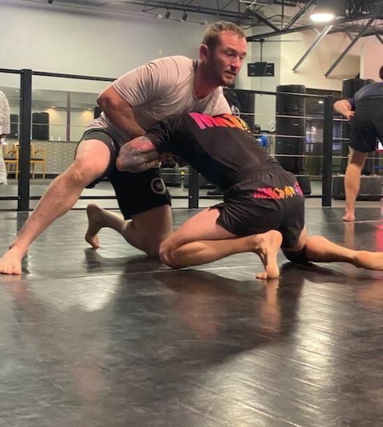 Two men grappling on a gym floor; one stands over the other in a wrestling or Jiu-Jitsu practice.