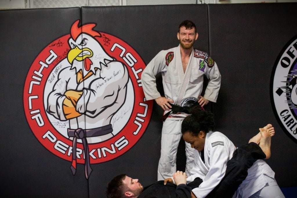 Three people in a Jiu-Jitsu gym. Two are grappling on the mat while a third stands with a black belt.