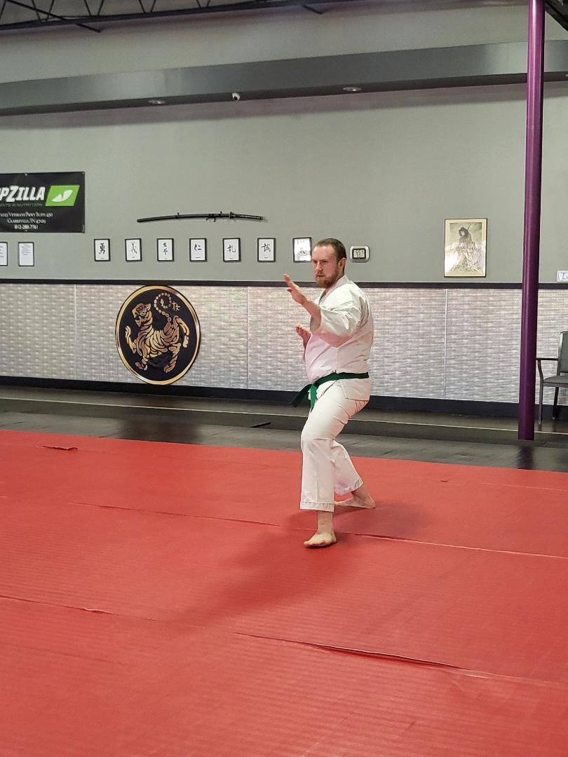 Man in karate uniform, green belt, executing a punch on a red mat in a dojo.
