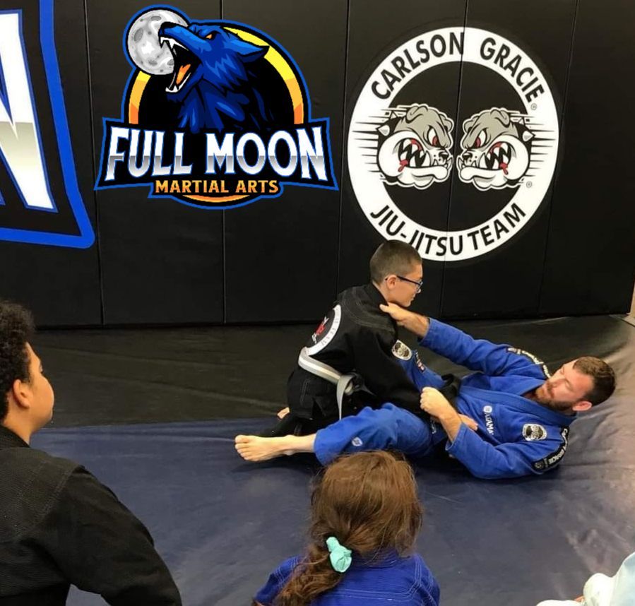 Two people practicing Jiu-Jitsu on a mat in a gym with logos. One in black gi, choking another in blue gi. Others observe.
